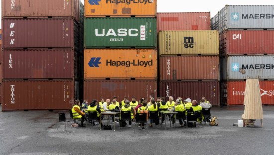 bilde av en gjeng som sitter i en sirkel blant kontainere på Dokken - Arild Eriksen "Hva vi lagde ved sjøen". Foto: Thor Brødreskift