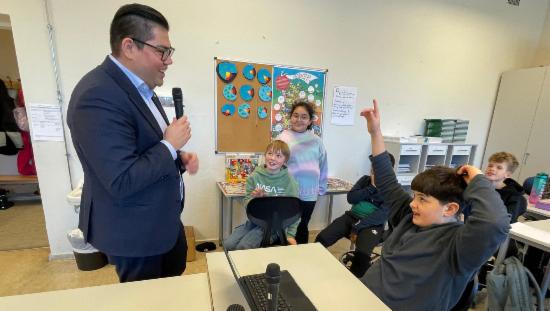 Mann i dress snakker i mikrofon med elever i et klasserom.