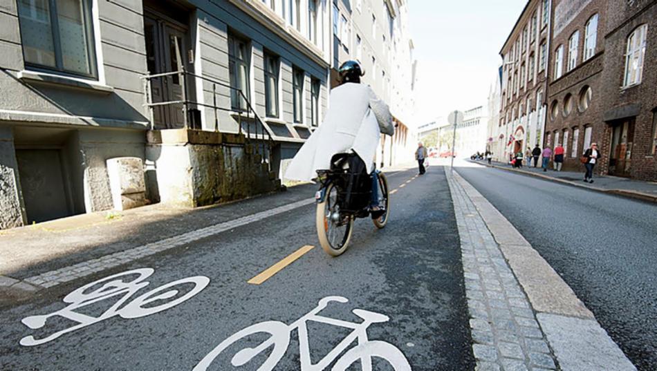 Syklist  i hvit frakk og hjelm og med sykkelveske på høyre bakhjul i asfaltert sykkelfelt med hvite sykler malt i begge retninger.Hus å begge sider og vei mellom