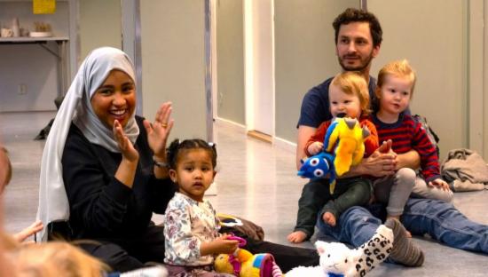 Dame med hijab med barn på fanget og mann med to barn på fanget sitter på gulvet 